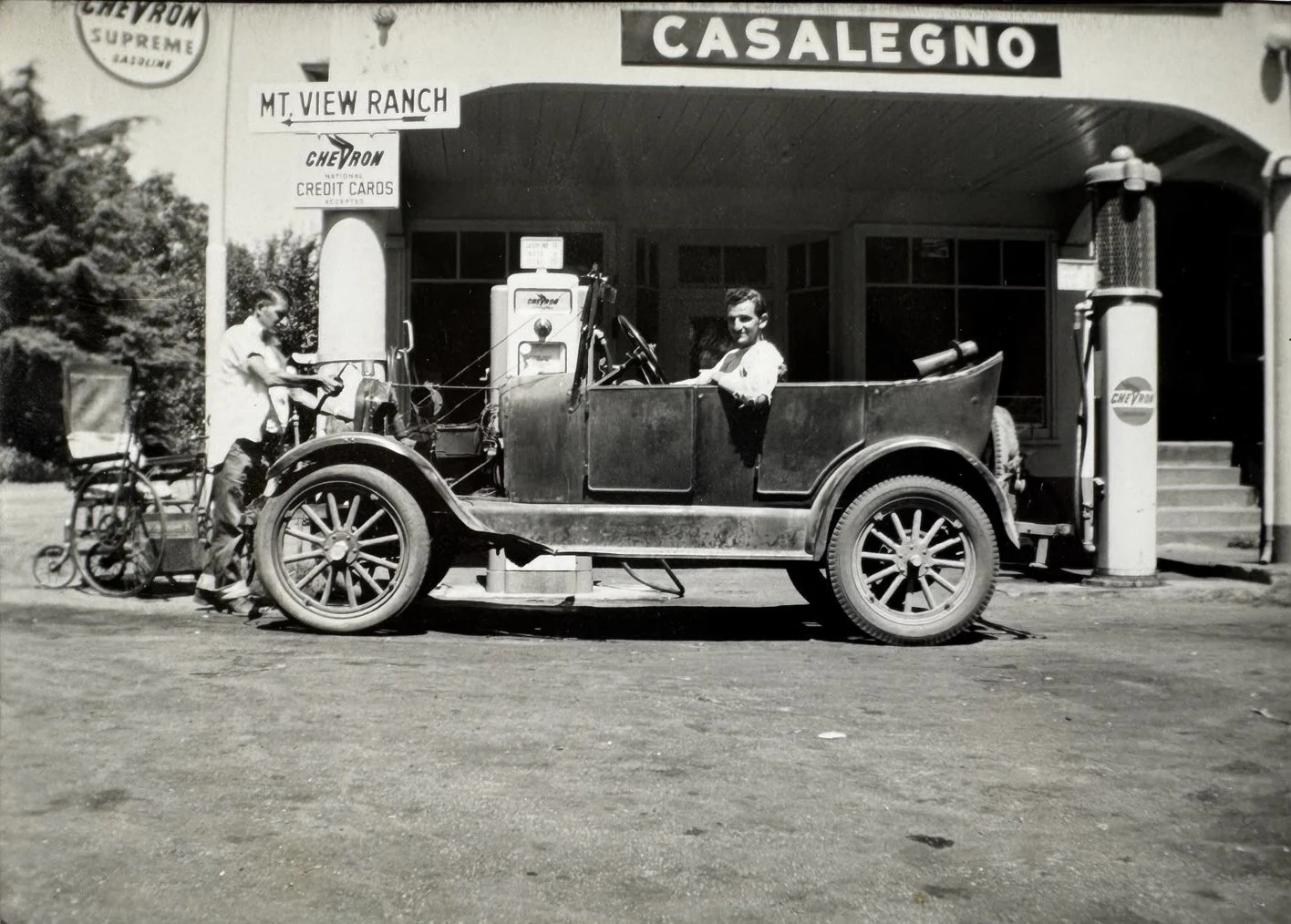 Black and white photo of a vintage Chevrolet pickup truck parked in front of a service station with signs for Chevron and Mount View Ranch. Two men are present; one standing near a bicycle and the other sitting in the truck's cab, smiling at the camera.