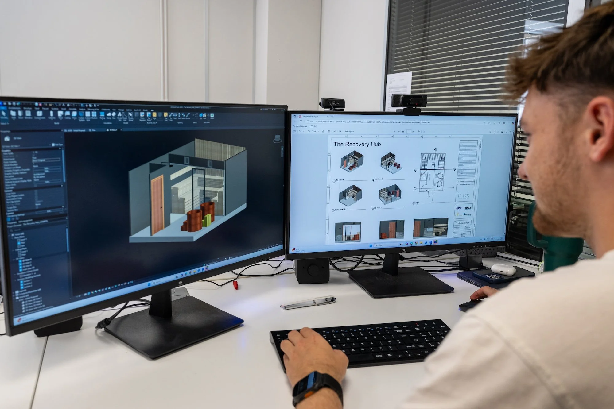 Person working on dual computer monitors displaying architectural design plans and 3D models in an office environment.
