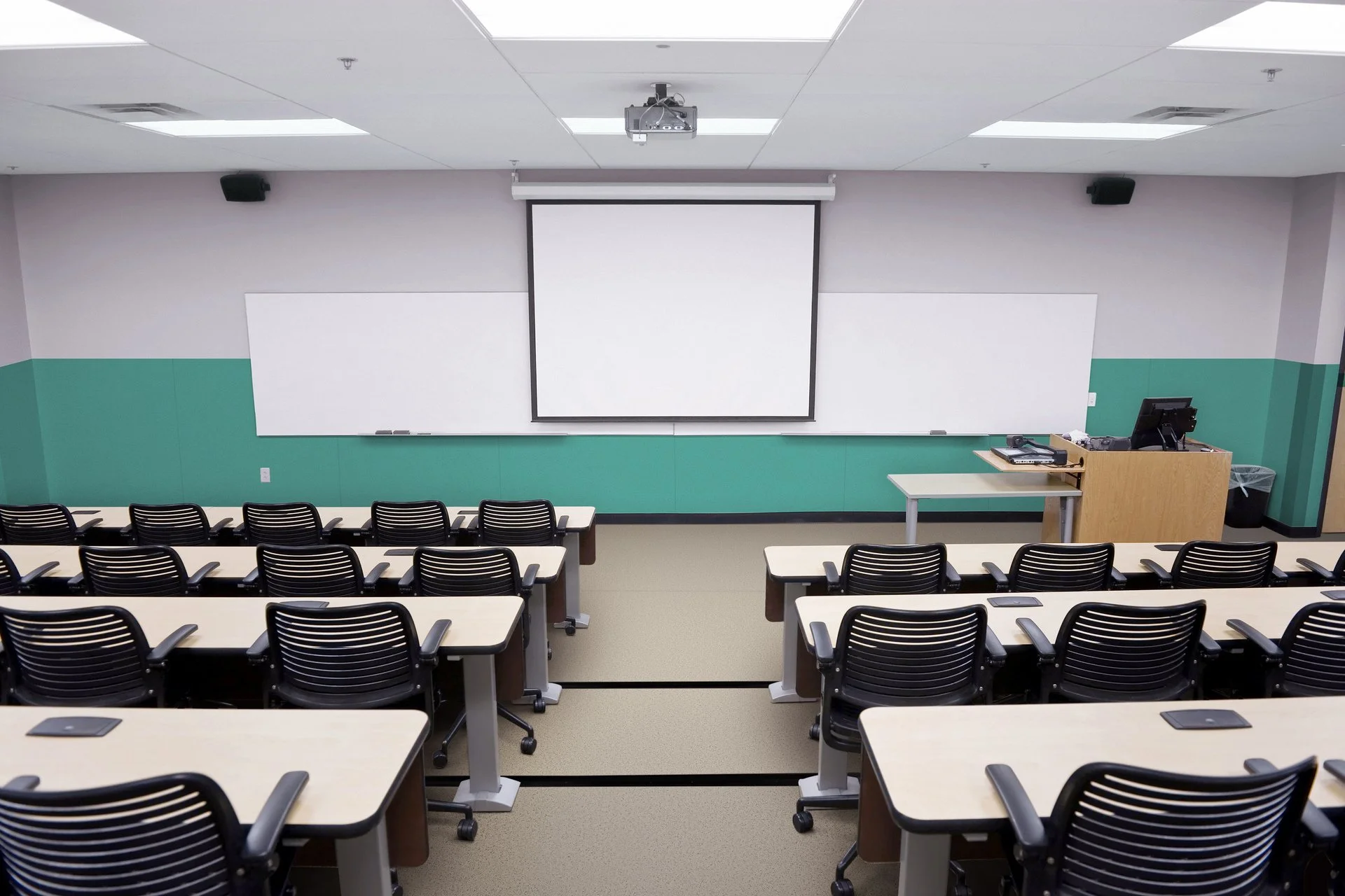 Empty classroom with desks, chairs, a whiteboard, a projection screen, and a teacher's desk with a computer, microphone, and trash can.