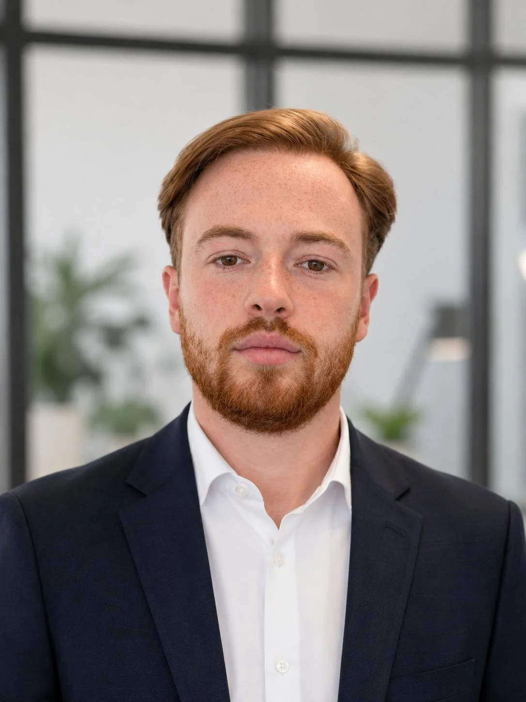 A man with red hair and a beard wearing a dark suit and white shirt stands indoors with a blurred office background.