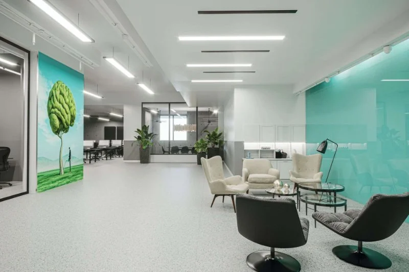 Modern office lounge area with armchairs, a floor lamp, glass coffee tables, and teal accent wall, with a large art piece of a tree and two figures.
