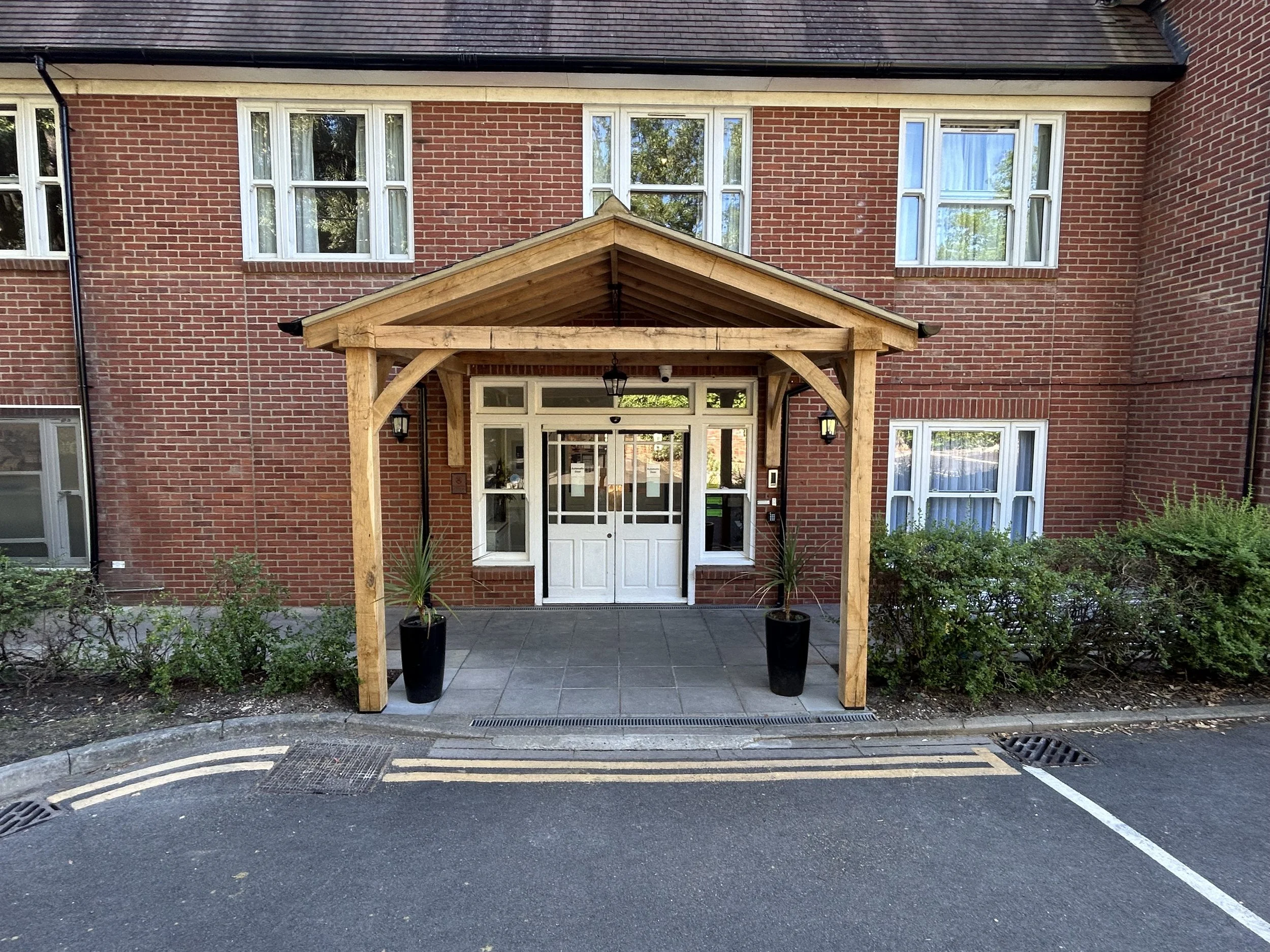 Entrance of a red brick building with large windows and a wooden canopy over the door, surrounded by greenery and a paved sidewalk.
