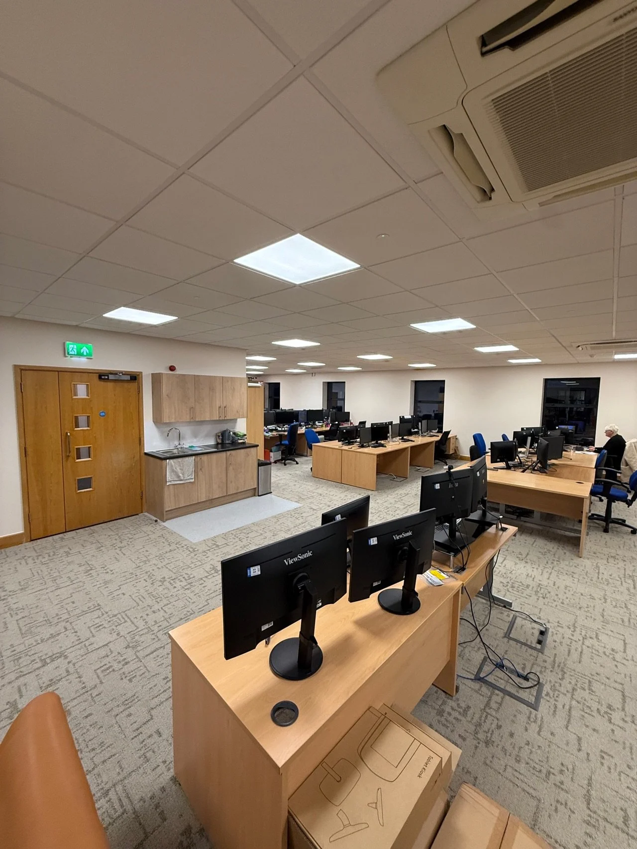 An office with multiple desks and computer monitors, a kitchenette with a wooden cabinet and sink, and a few people working at their desks. The ceiling has square light panels and an air conditioning vent, and the floor is carpeted.