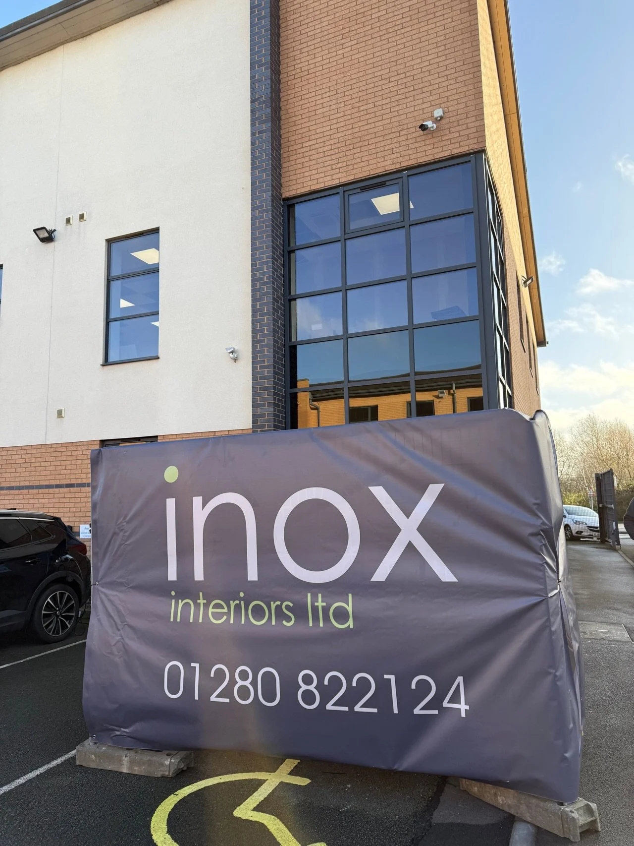 A building with white and brick walls featuring a large multi-pane window. In the foreground, there is a construction fence banner displaying 'Inox Interiors Ltd' and a phone number.