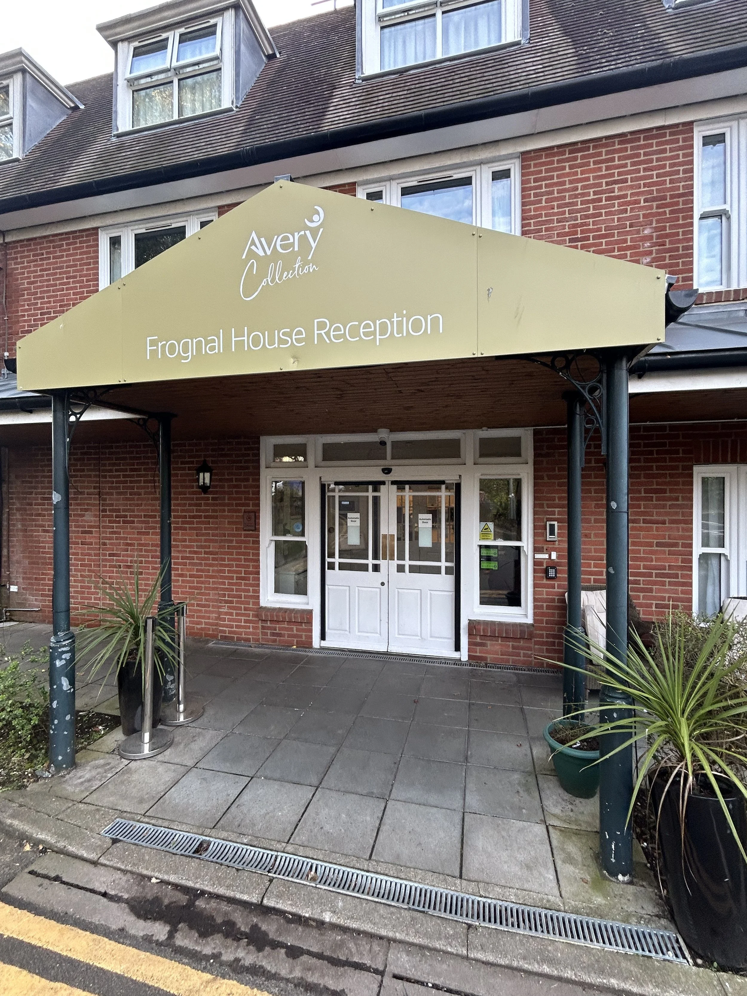 Sign reading 'Avery Collection Frogmal House Reception' outside a brick building with windows and an entrance, decorated with potted plants and a textured walkway.