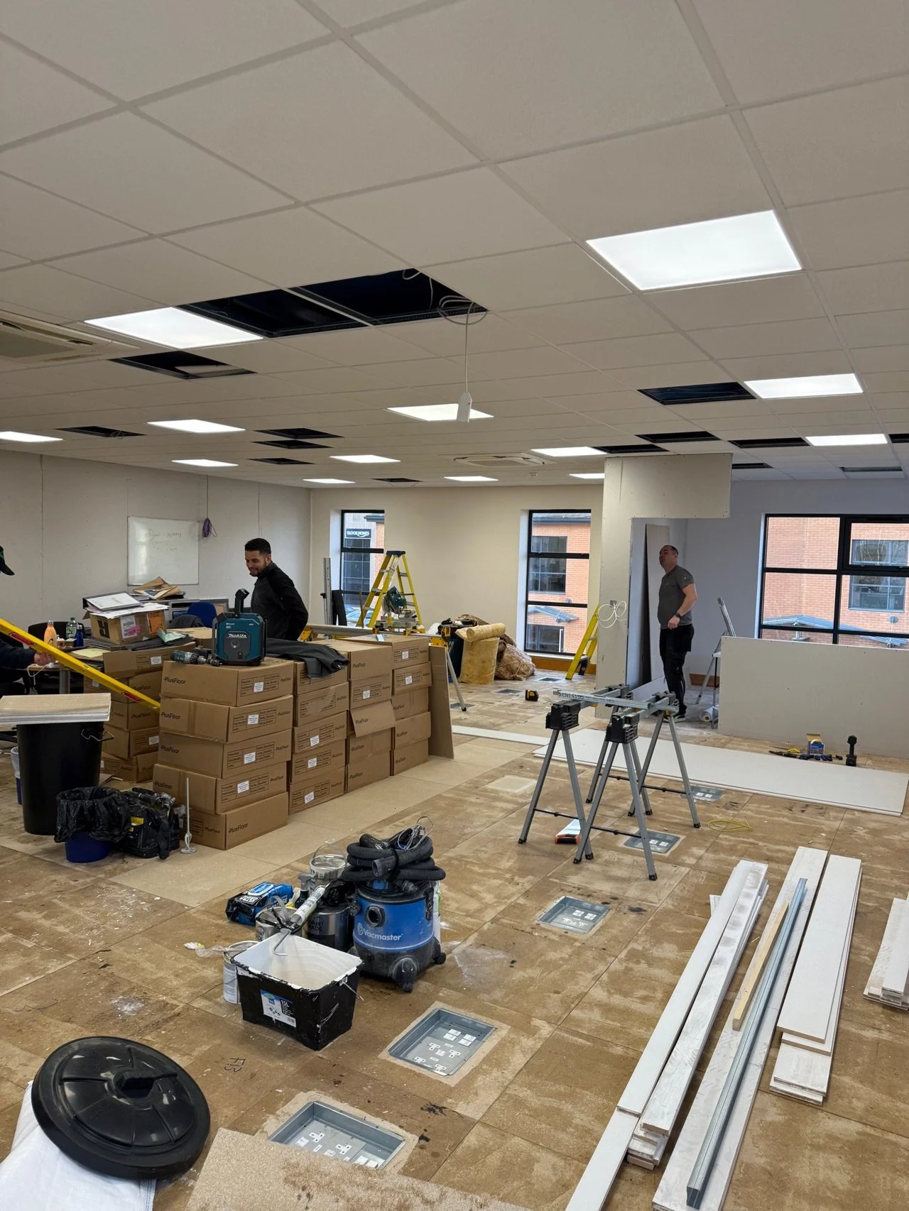 Interior of a building under renovation with construction workers, tools, boxes, and partially installed ceiling panels.