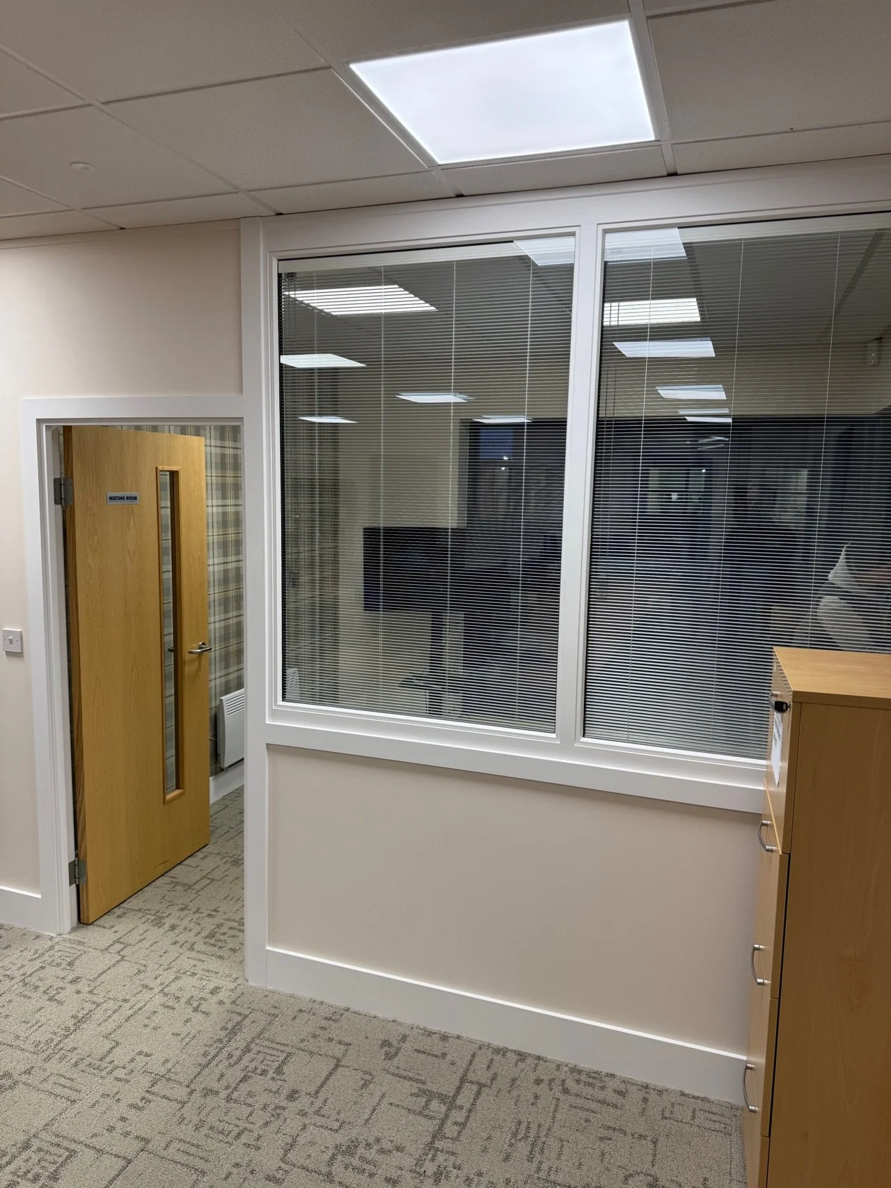 Interior view of an office with a wooden door labeled 'Meeting Room,' large glass windows with blinds, a TV, and a wooden cabinet.