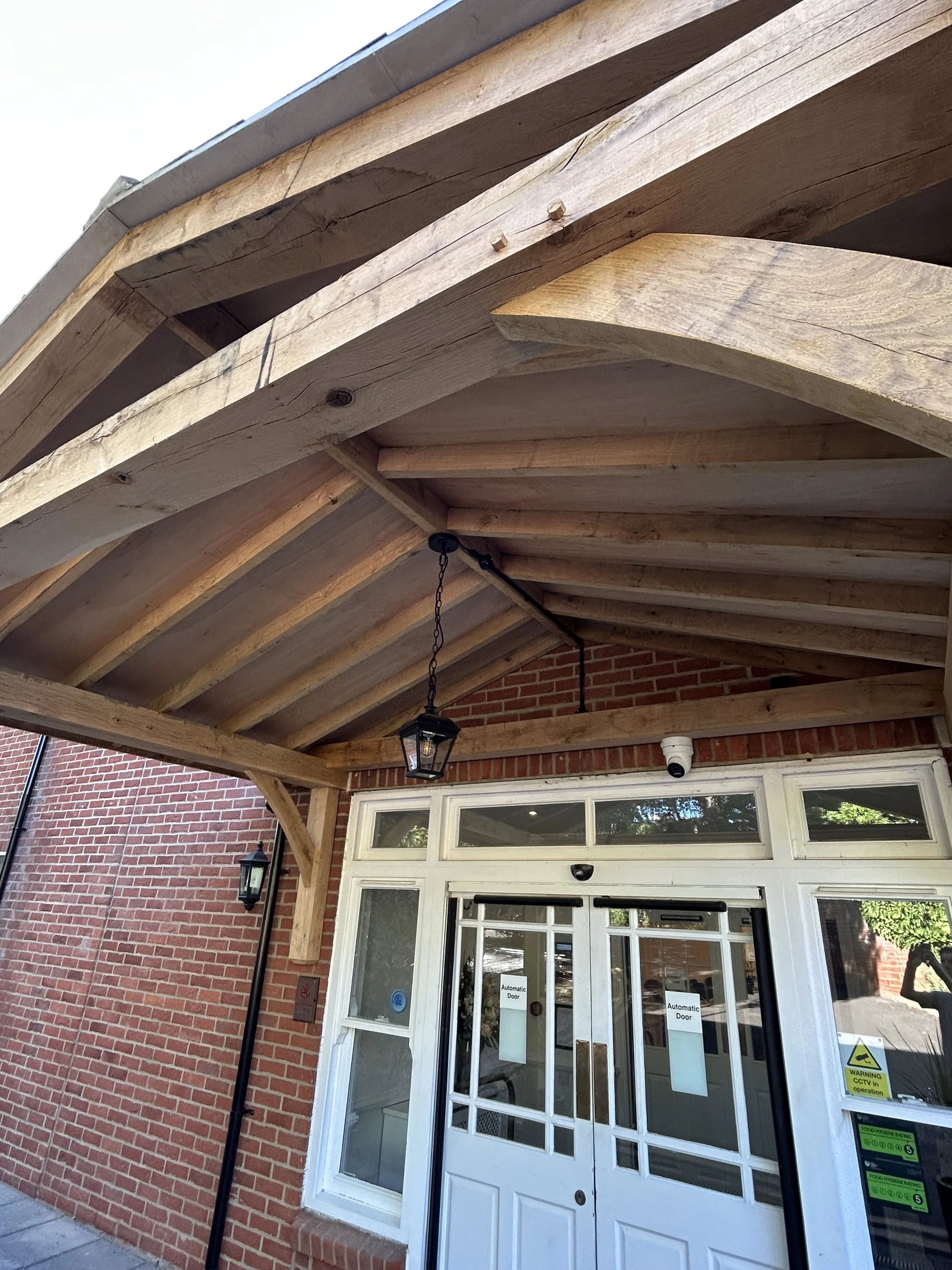 Exterior of a brick building with glass doors labeled 'Automatic Door,' hanging lantern lights, a security camera, and a wooden canopy structure above.
