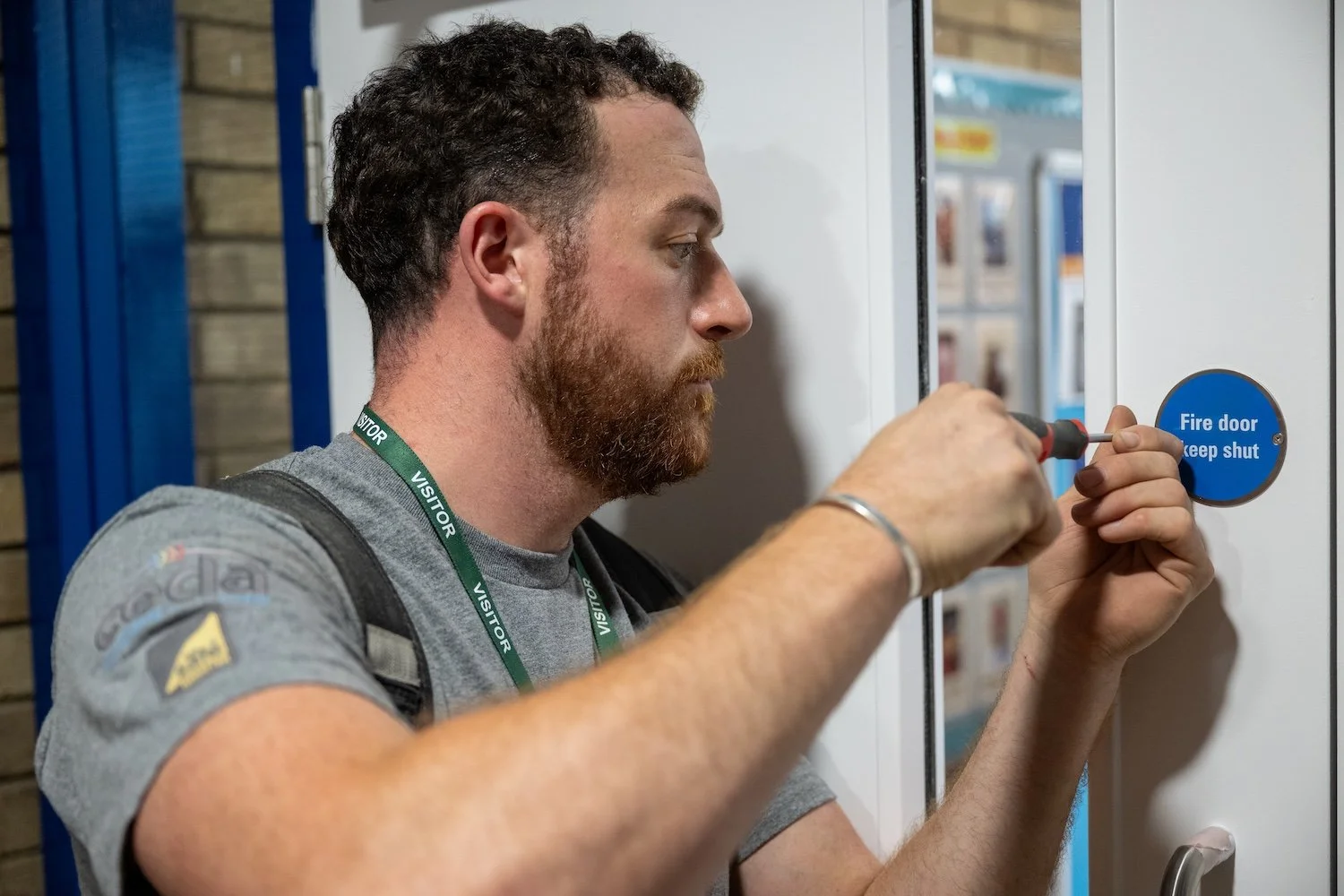A man with a beard and a backpack fixing a blue circular sign that says 'Fire door keep shut' on a white wall.