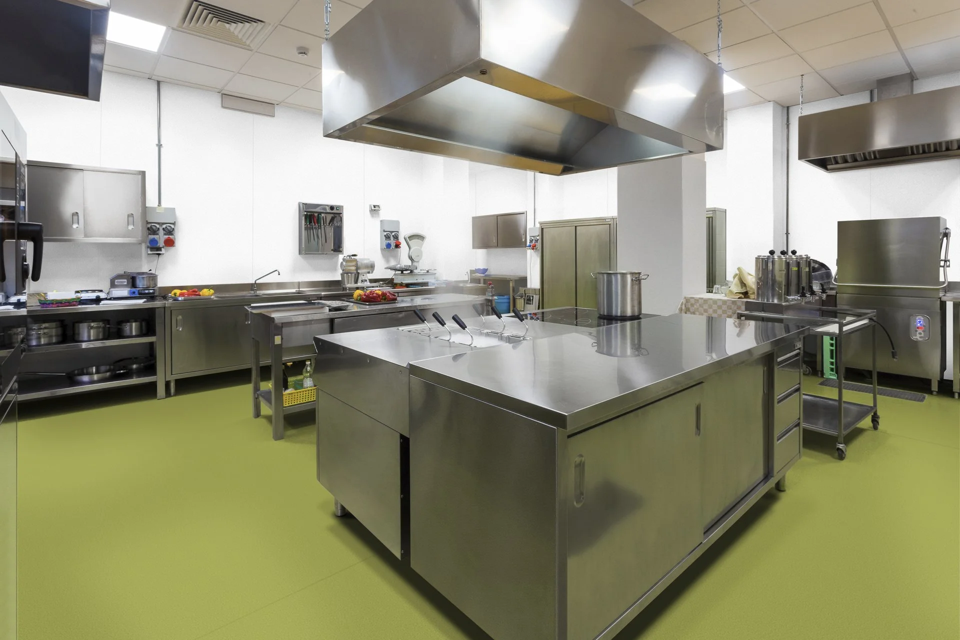 A clean, professional commercial kitchen with stainless steel appliances and workstations, green flooring, and white walls.
