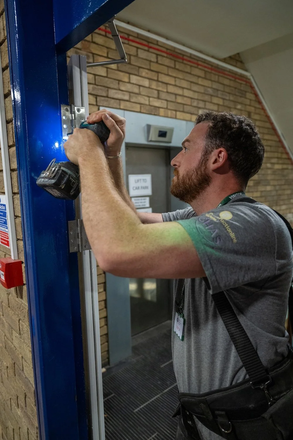 Man installing or repairing a metal frame with a power drill, in an indoor environment near a brick wall and an elevator door.