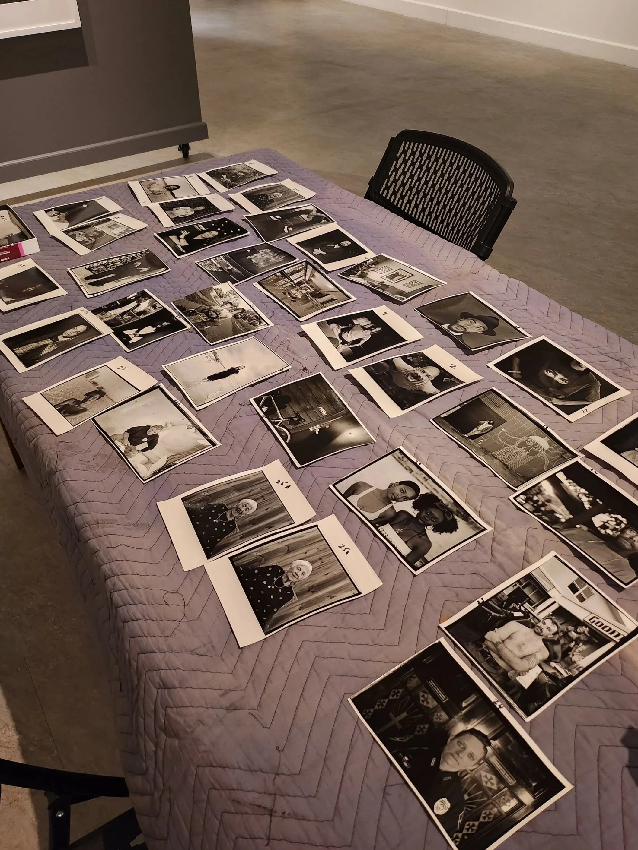 A table displaying multiple black-and-white photographs of various people, landscapes, and objects. A black chair is partially visible on the right side of the table.