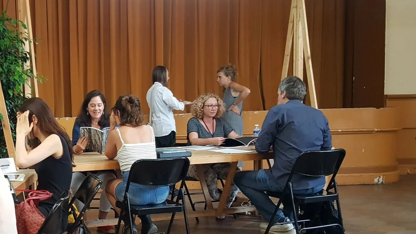 Group of people sitting and standing at a long table in a room with wooden walls, talking, or interacting and reviewing portfolios