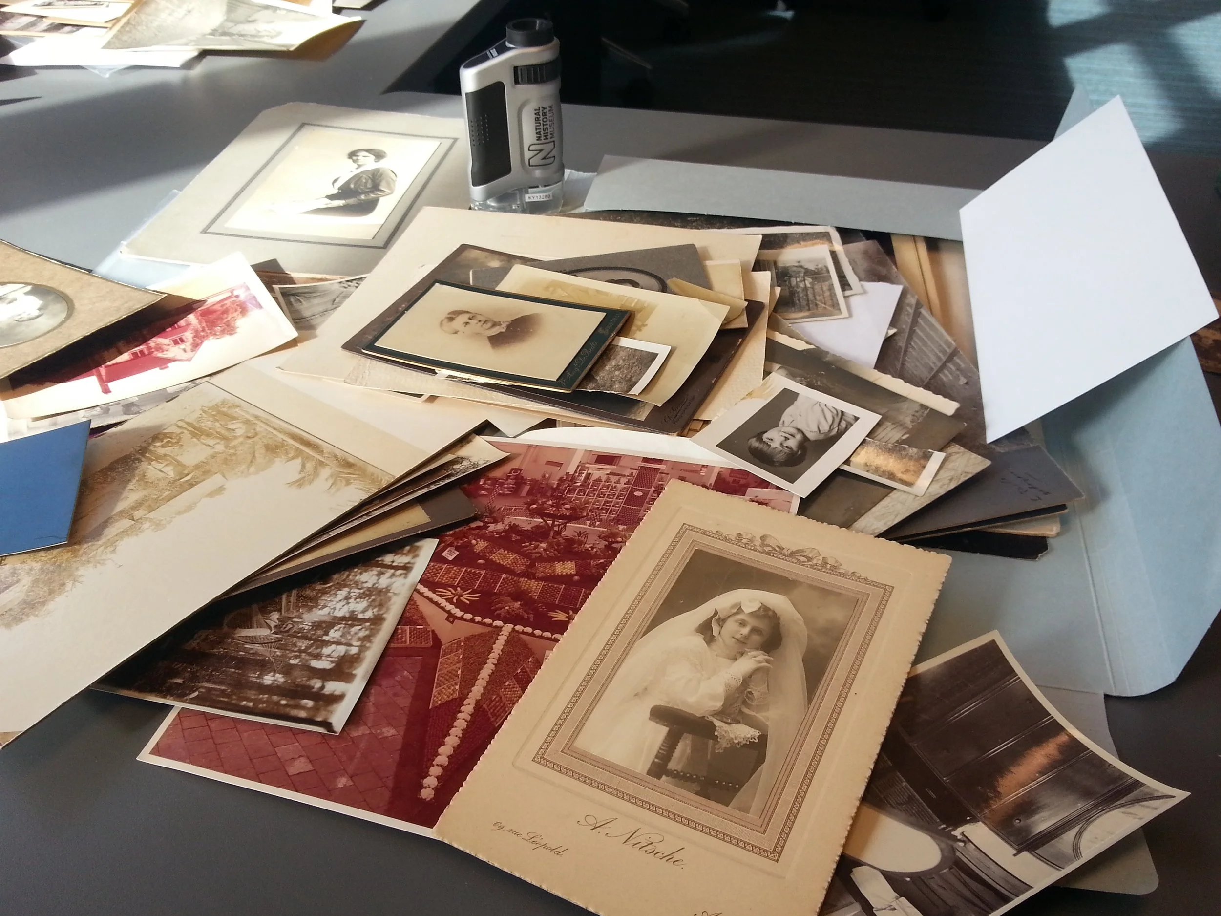A cluttered table with numerous antique photographs, postcards, and paper documents scattered across the surface. Some photos feature portraits of women, and there is a variety of paper sizes and colors, including sepia-toned and black-and-white images.