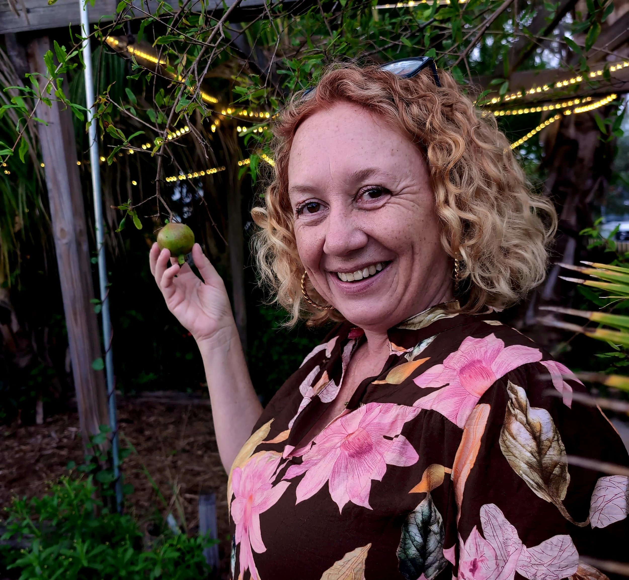 Woman with curly red hair smiling outdoors, holding a small green fruit, with greenery and string lights in the background.