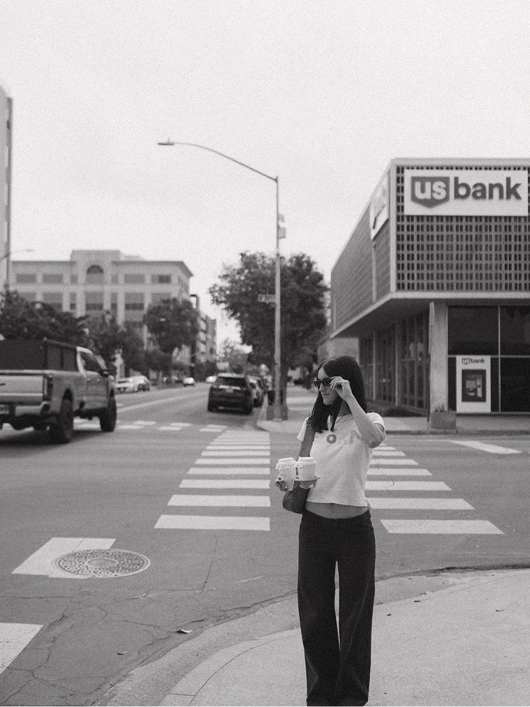 A woman standing at a crosswalk on a city street, holding takeout food and adjusting her sunglasses, with a US Bank building in the background.
