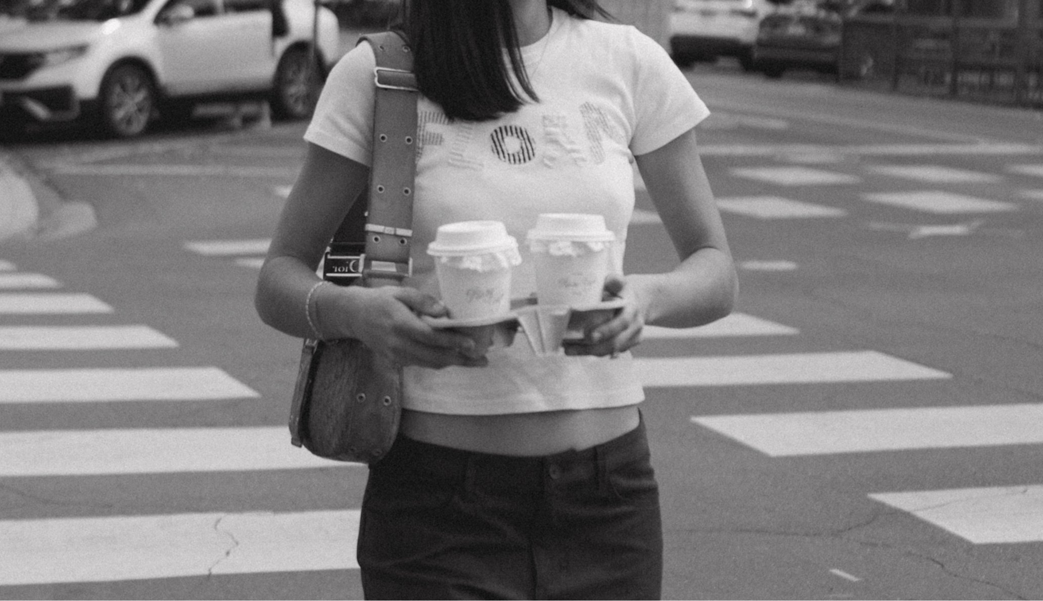 A woman walking across a crosswalk holding two coffee cups and a tray with two cups, wearing a t-shirt with the word 'FOOD' on it and a shoulder bag.