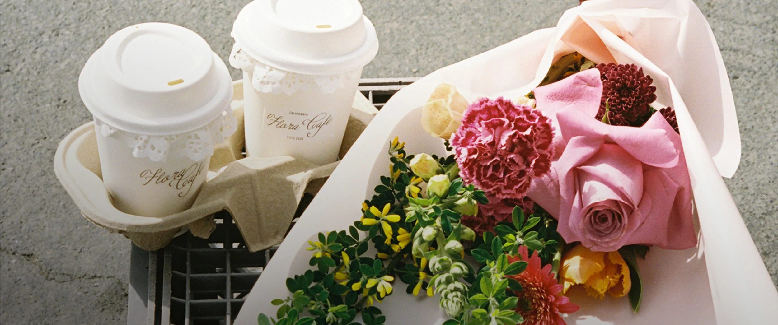 Two to-go coffee cups in a cardboard holder beside a bouquet of pink and red flowers on a sidewalk.