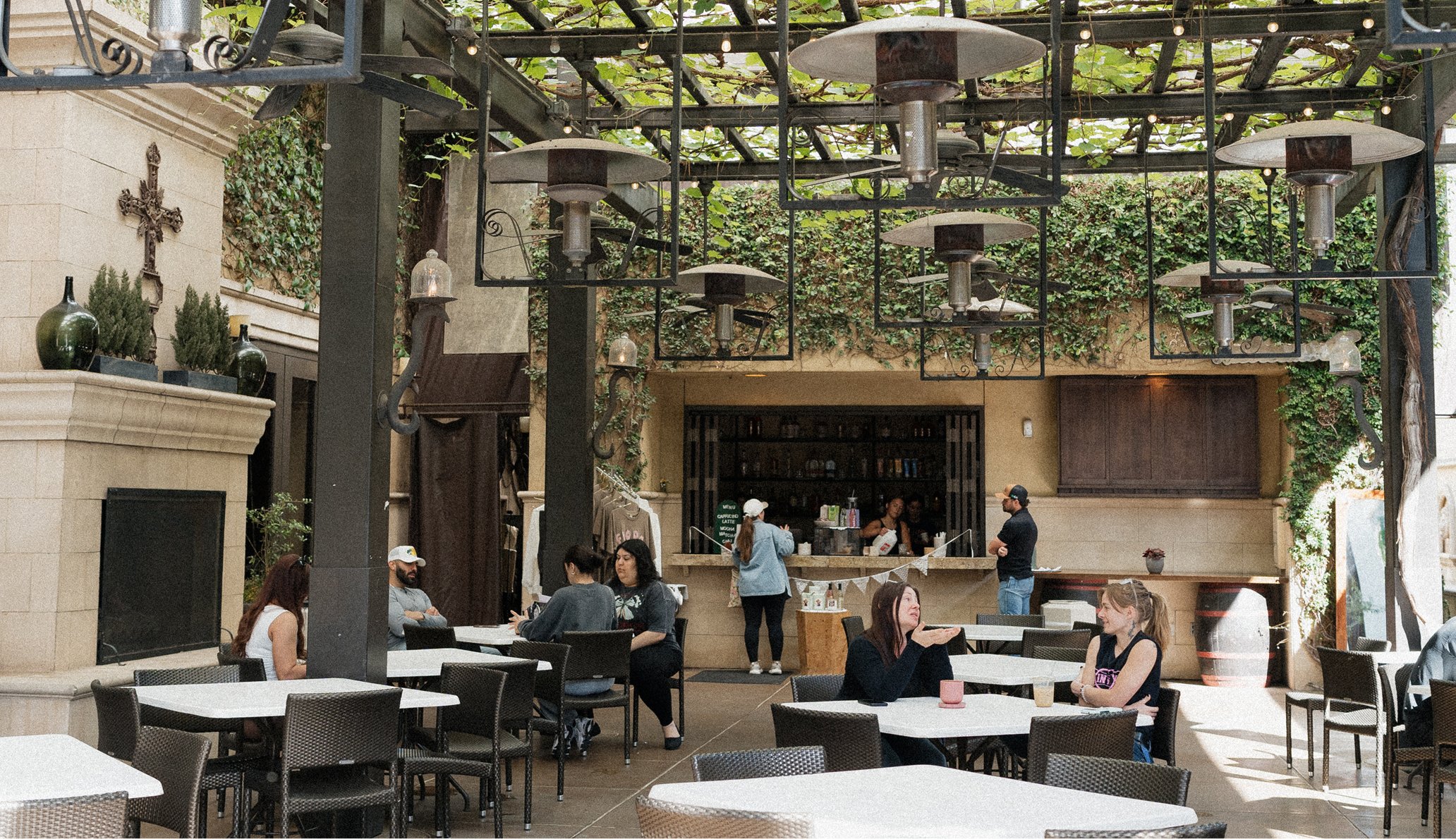 Outdoor restaurant patio area with tables and chairs, people dining, a coffee bar in the background, and hanging heaters under a trellis.