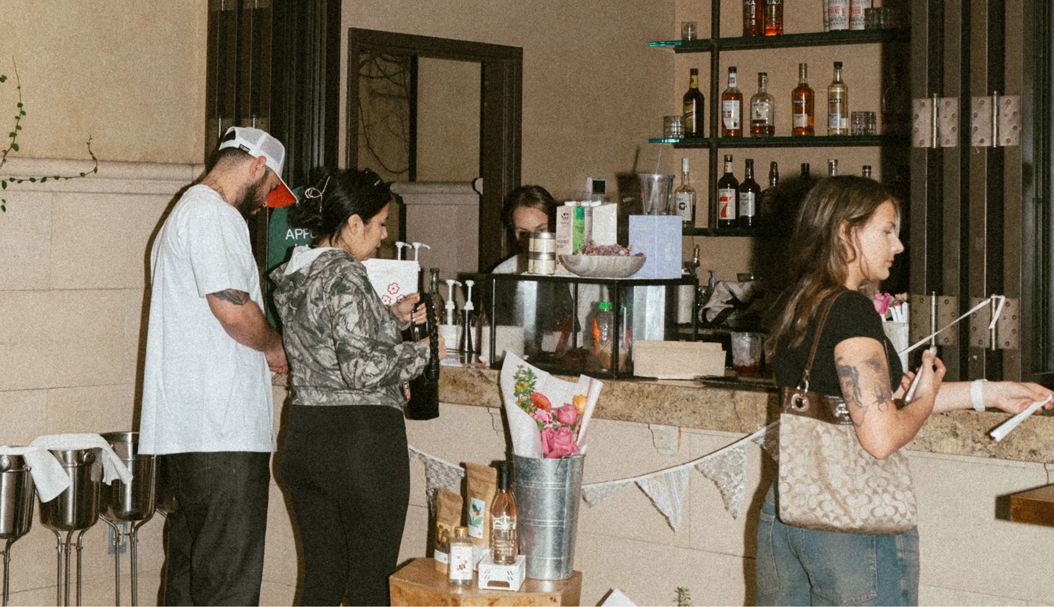 People at a bar counter, with alcohol bottles on shelves behind them, one person holding a smartphone, another person filling a glass, and a third woman reading a menu or taking an order.