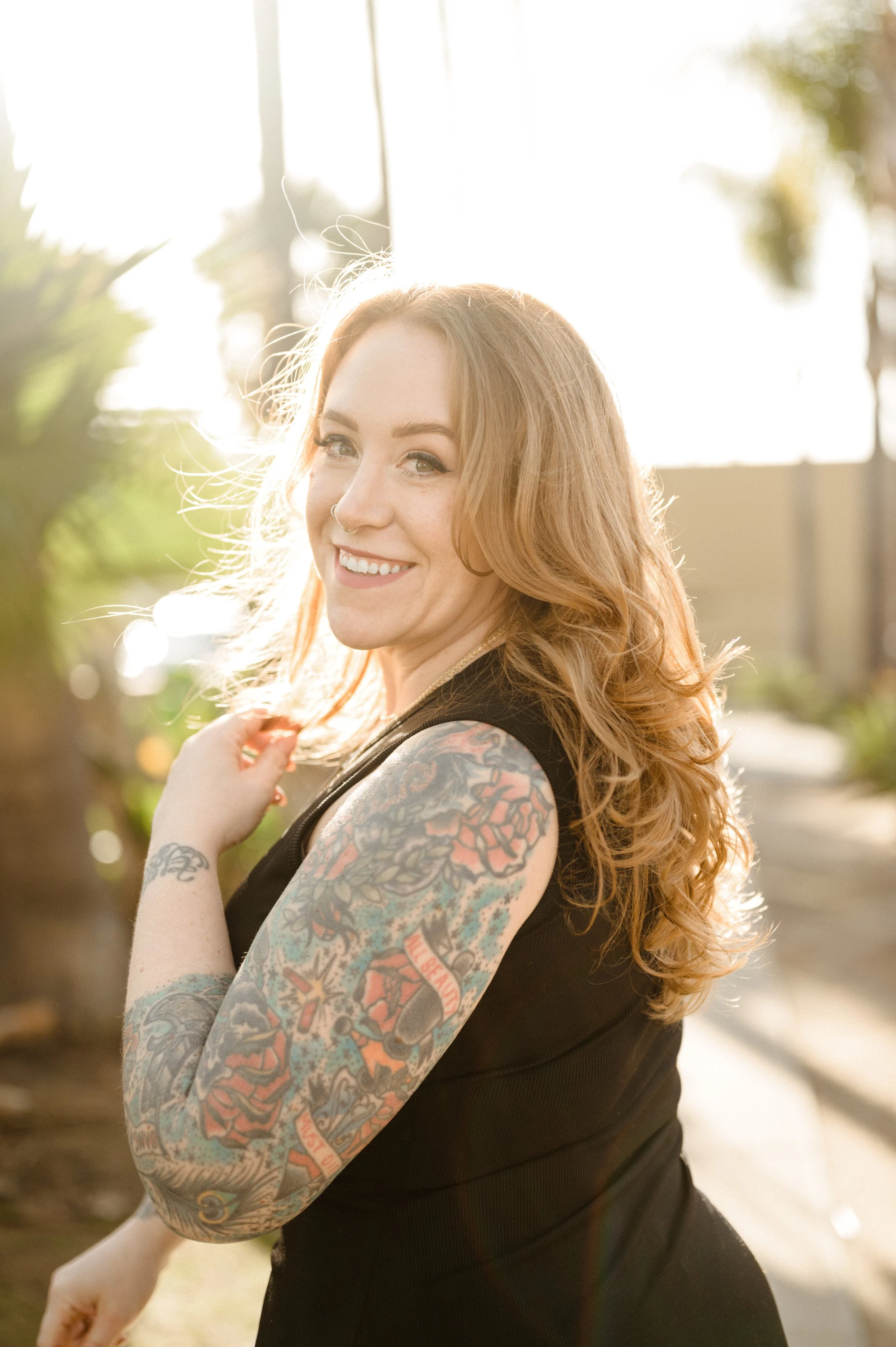 A woman with long, curly red hair smiling outdoors in sunlight, showing colorful tattoos on her arms, wearing a black sleeveless top.