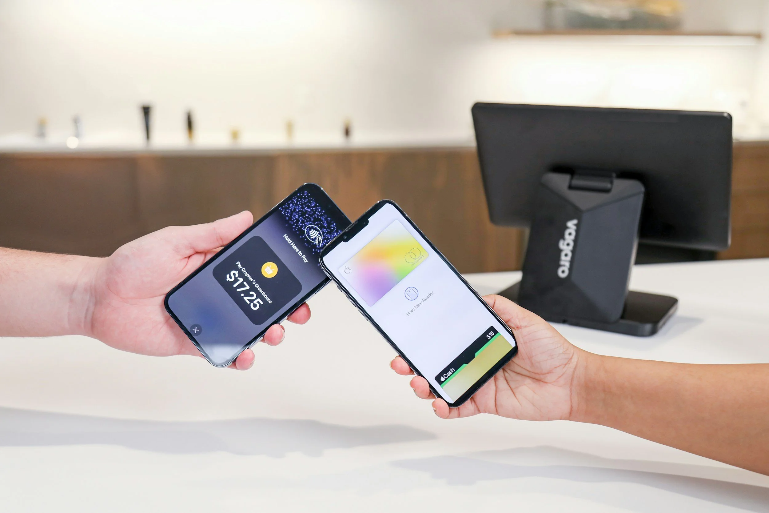 Person holding two smartphones with digital payment screens at checkout counter with a card reader and countertop.