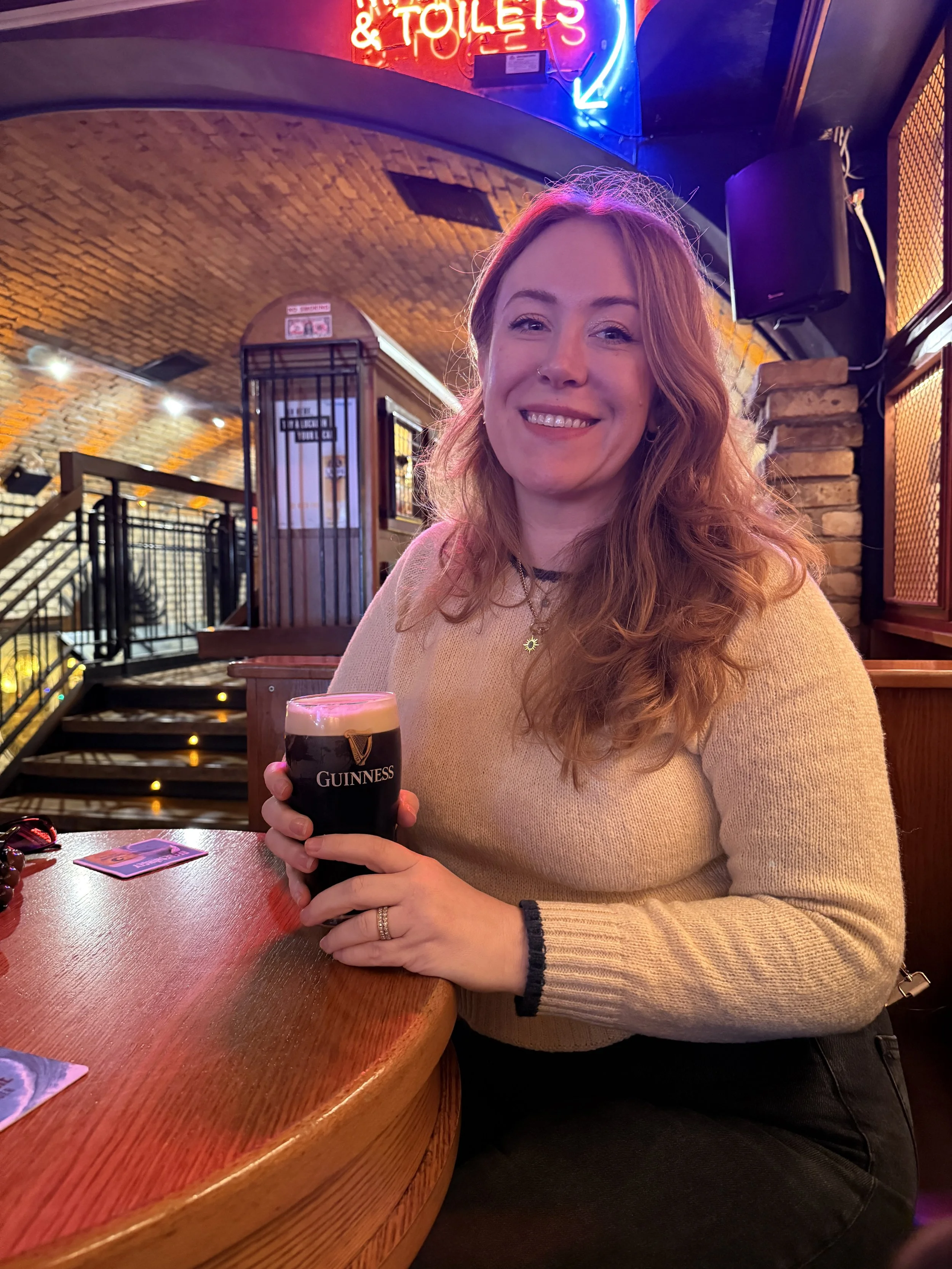 A woman sitting at a wooden bar in a dimly lit pub, holding a pint of Guinness beer with a smile. Neon signs and brick walls in the background.