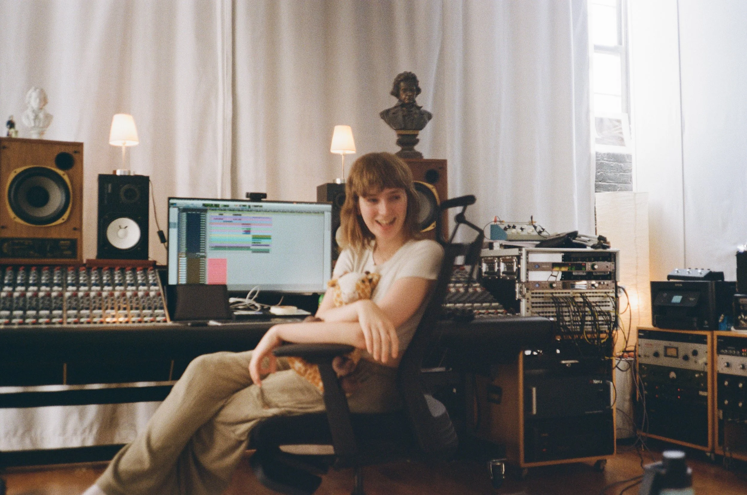 Jamie sits in a recording studio with a smile, surrounded by music equipment including speakers, a computer with audio editing software, and various audio gear.