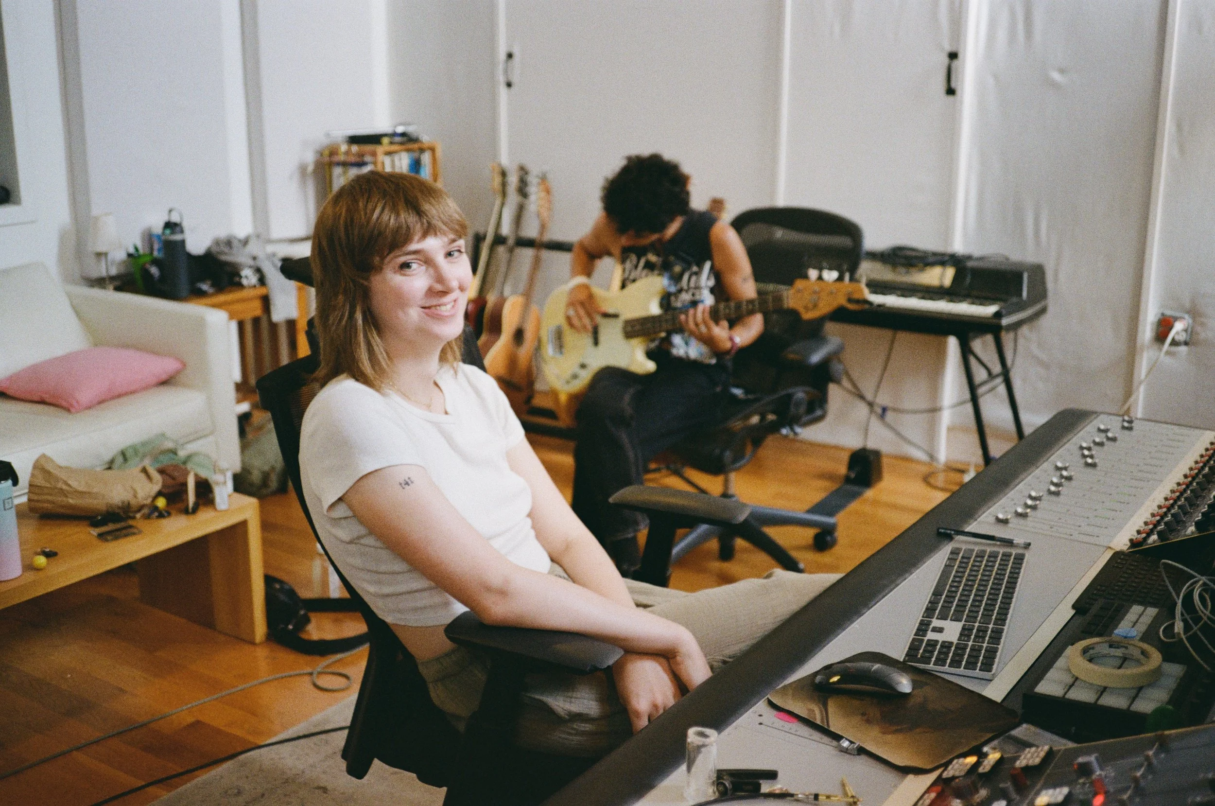 Jamie smiling at the camera in a music studio surrounded by musical instruments and equipment.
