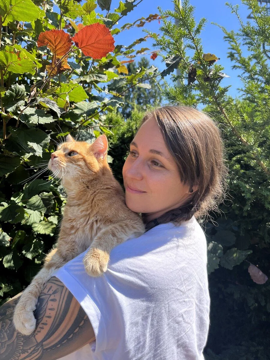 Eine junge Frau hält eine orange Katze im Freien, umgeben von grünen Blättern und Bäumen bei sonnigem Himmel.