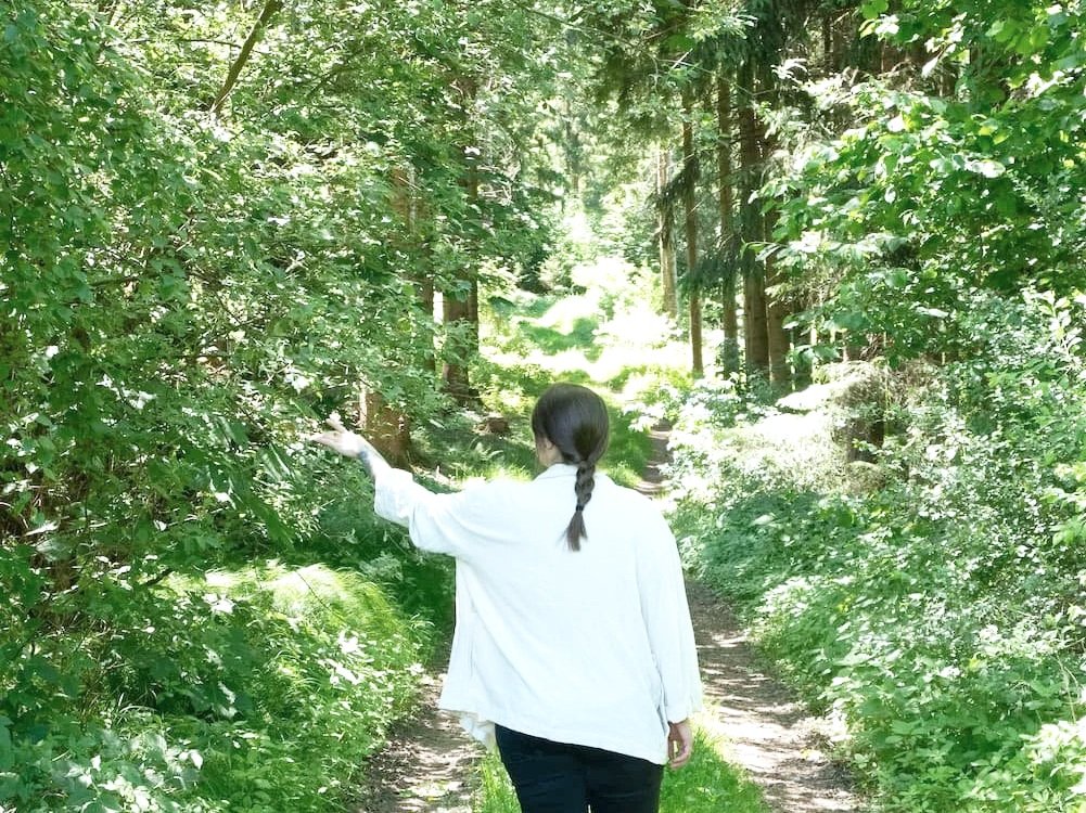Eine Person mit braunem Pferdeschwanz trägt ein weißes Hemd und geht einen Waldweg entlang, umgeben von grünen Büschen und Bäumen.