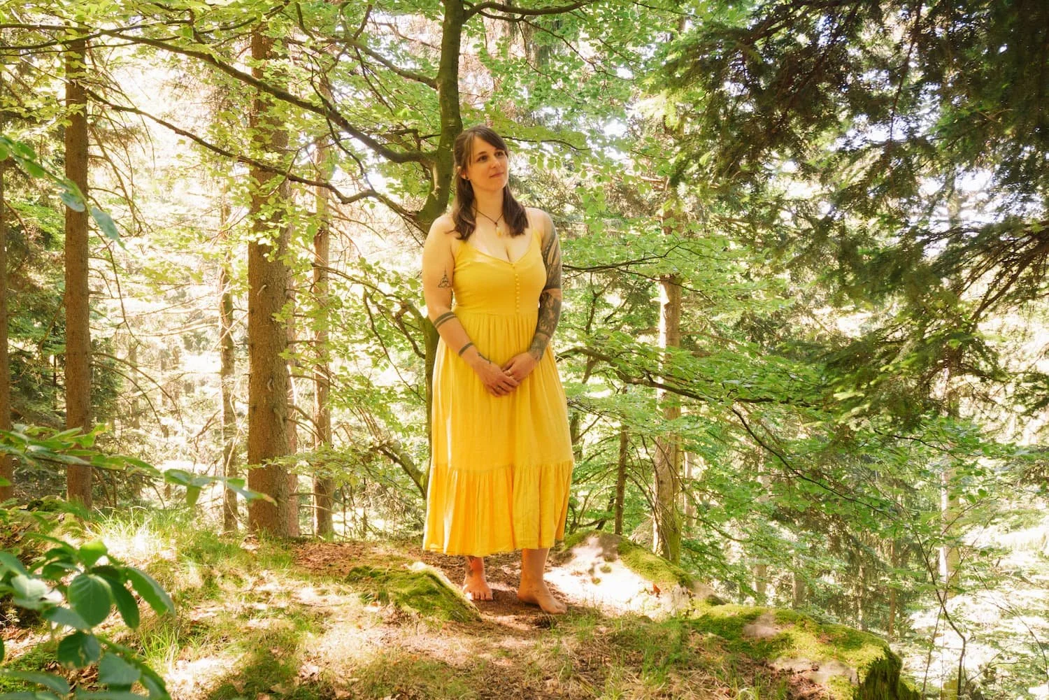 Eine Frau in einem gelben Kleid steht barfuß auf einem Baumstamm in einem bewaldeten Gebiet, umgeben von grünen Bäumen und Sonnenlicht. Sie hat dunkles Haar und sichtbare Tätowierungen auf den Armen.