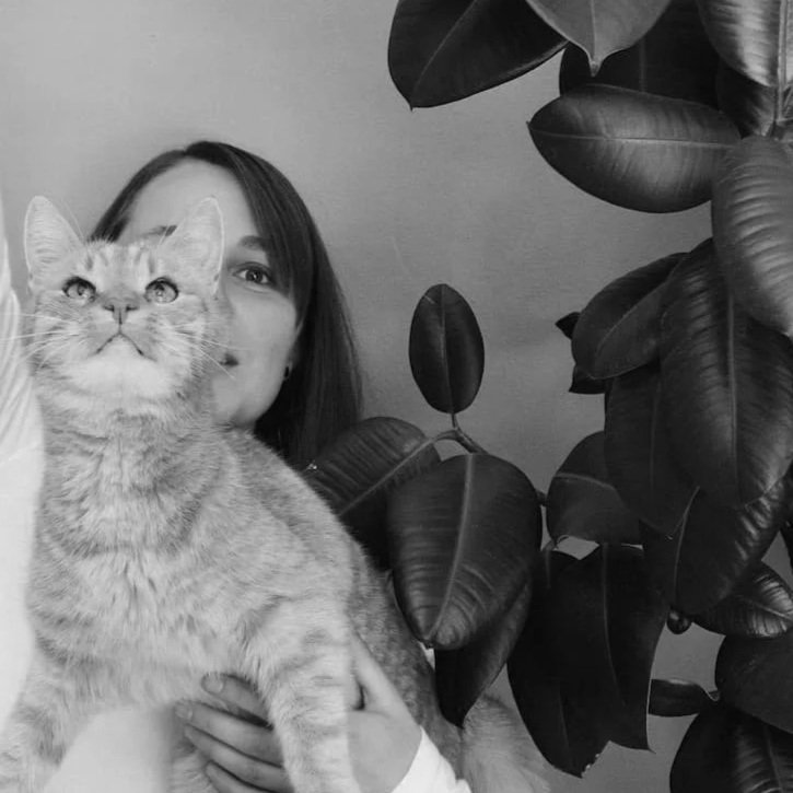 Ein Mädchen hält eine Katze vor ihrem Gesicht, im Hintergrund große dunkle Blätter einer Pflanze.