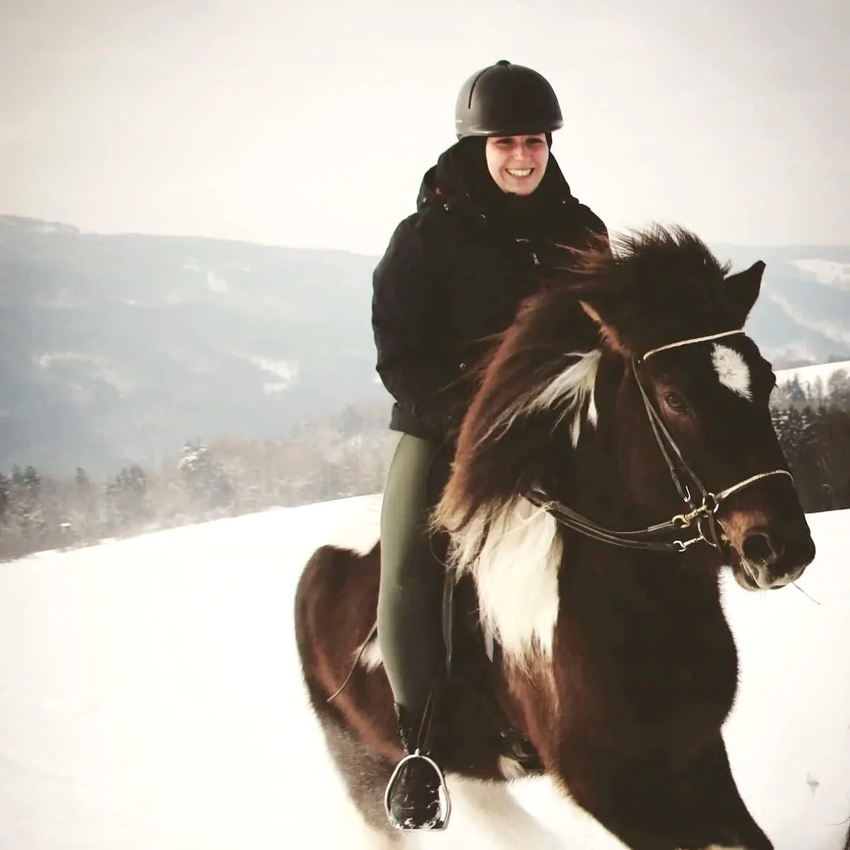 Junge Frau reitet im Winter auf einem Pferd in einer schneebedeckten Landschaft, trägt einen Reithelm und lächelt.