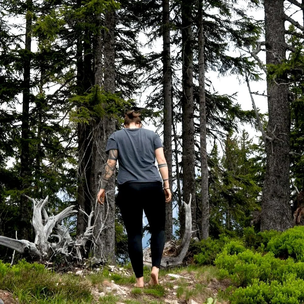 Eine Person mit Tattoos trägt eine graue T-Shirt und schwarze Hosen, läuft barfuß in einem Wald.