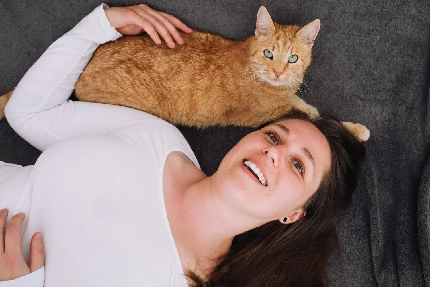 Eine lachende Frau mit braunen Haaren liegt auf einem schwarzen Sofa neben einer orangefarbenen Katze.