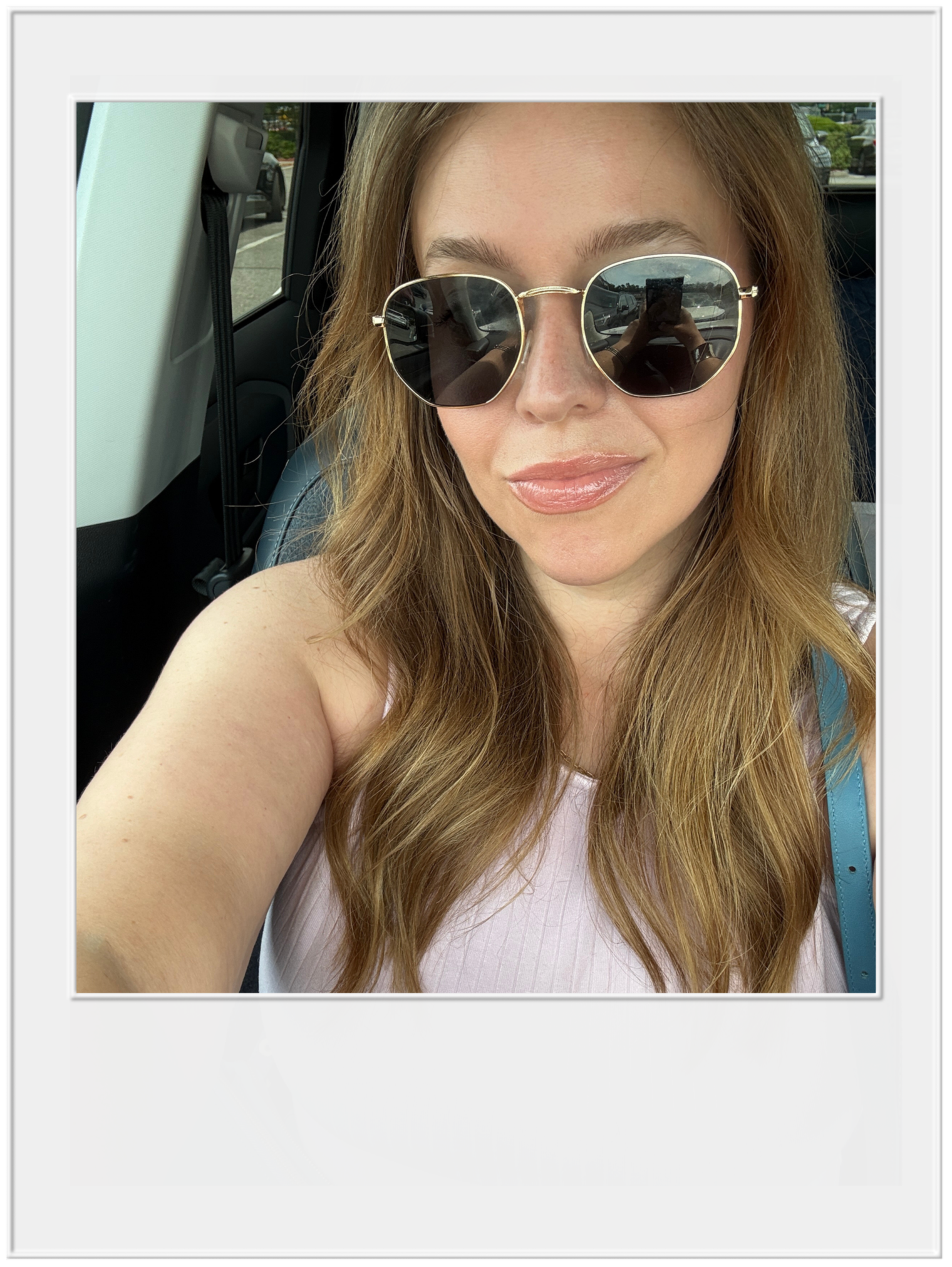 A woman with long red hair wearing reflective sunglasses, smiling slightly, sitting inside a vehicle.