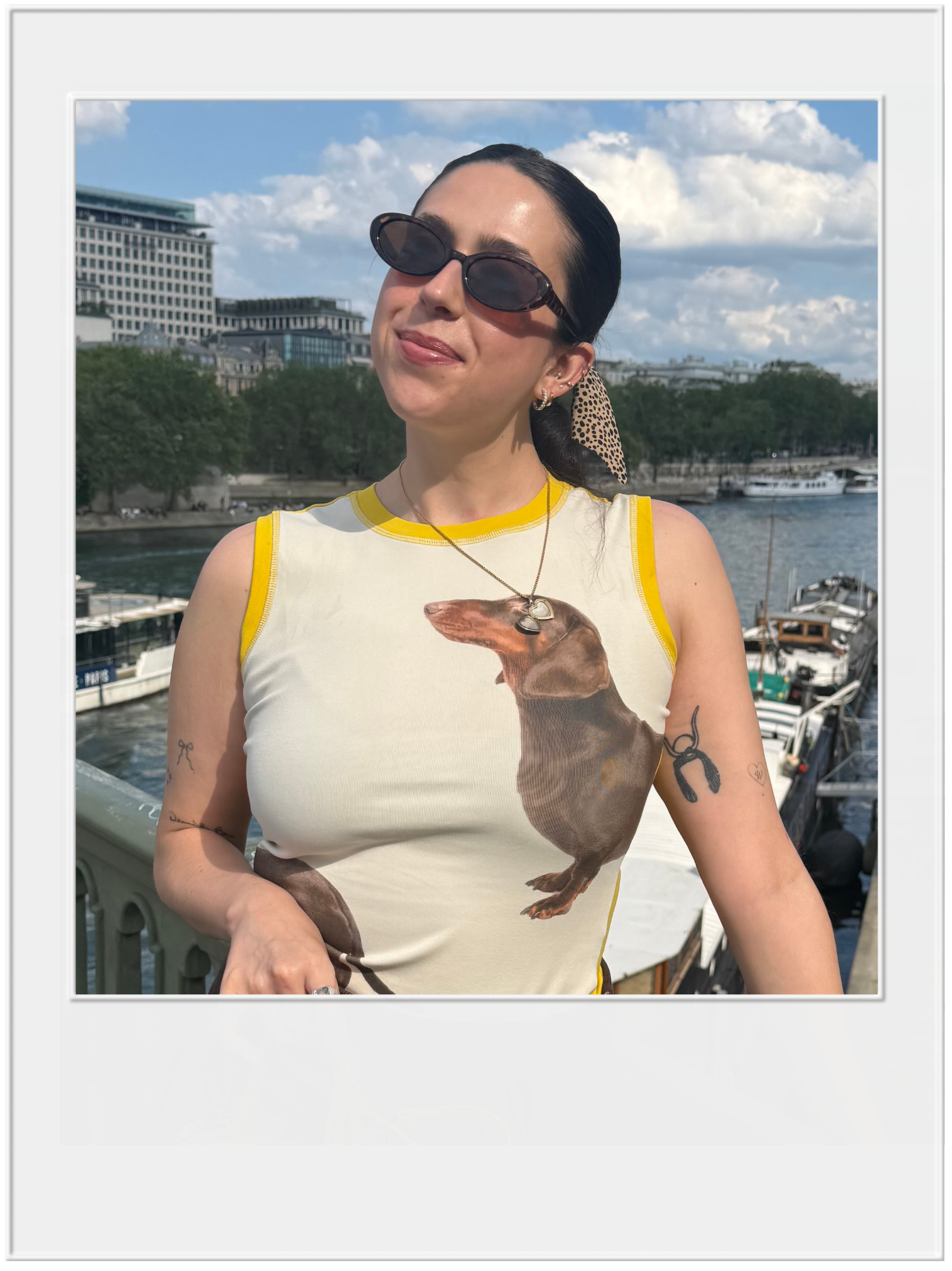 A woman with dark hair, wearing sunglasses and a sleeveless shirt with a dog print, standing outdoors with a cityscape and river in the background.
