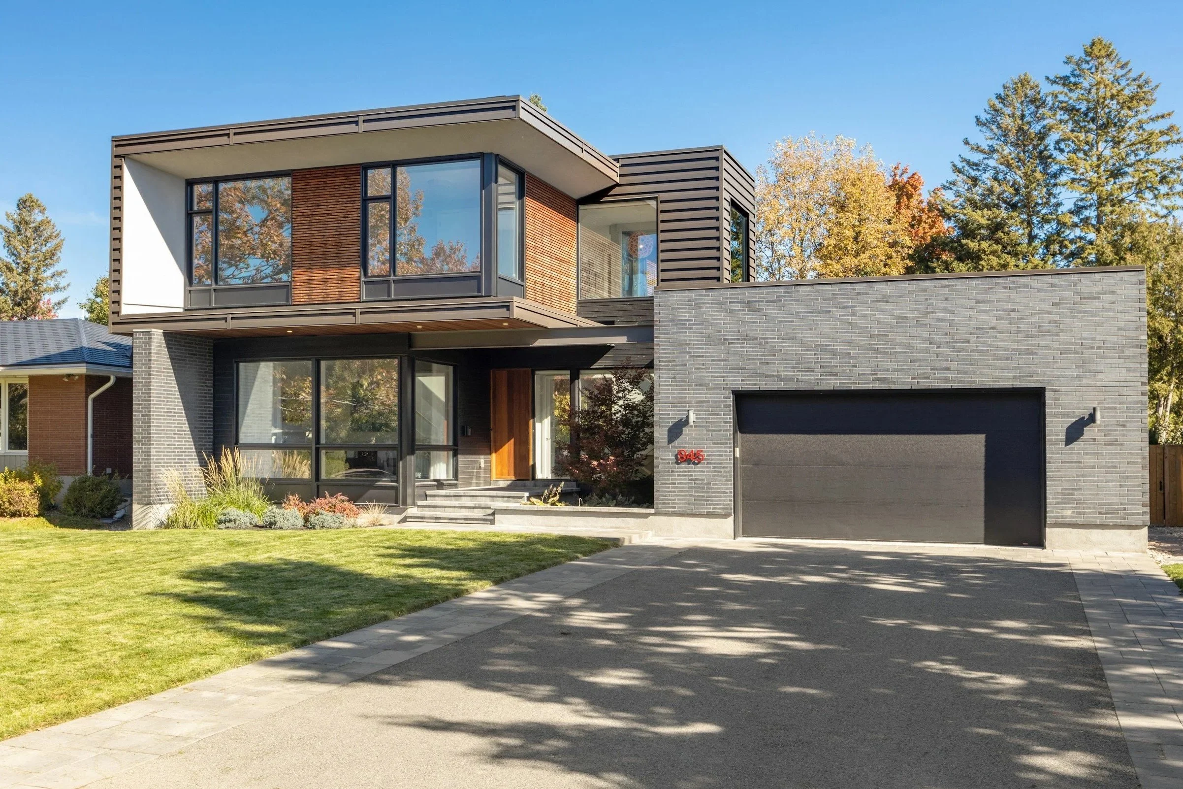 Modern two-story house with gray brick exterior, large windows, and wooden accents, surrounded by a well-maintained lawn and trees with fall foliage.