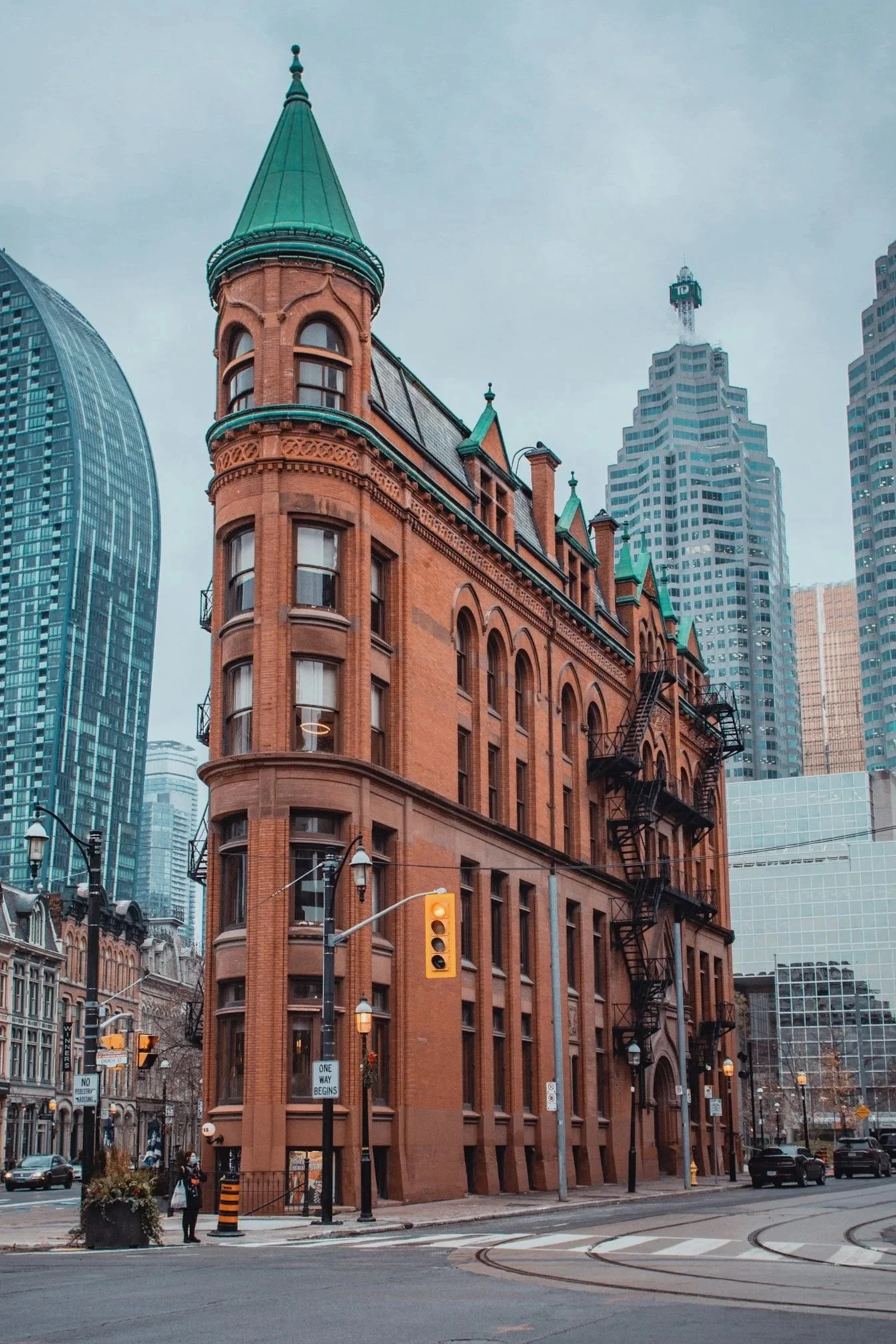 Historic red brick building with green roof and tower in an urban cityscape, featuring modern high-rise buildings in the background, street cars, traffic lights, and pedestrians.