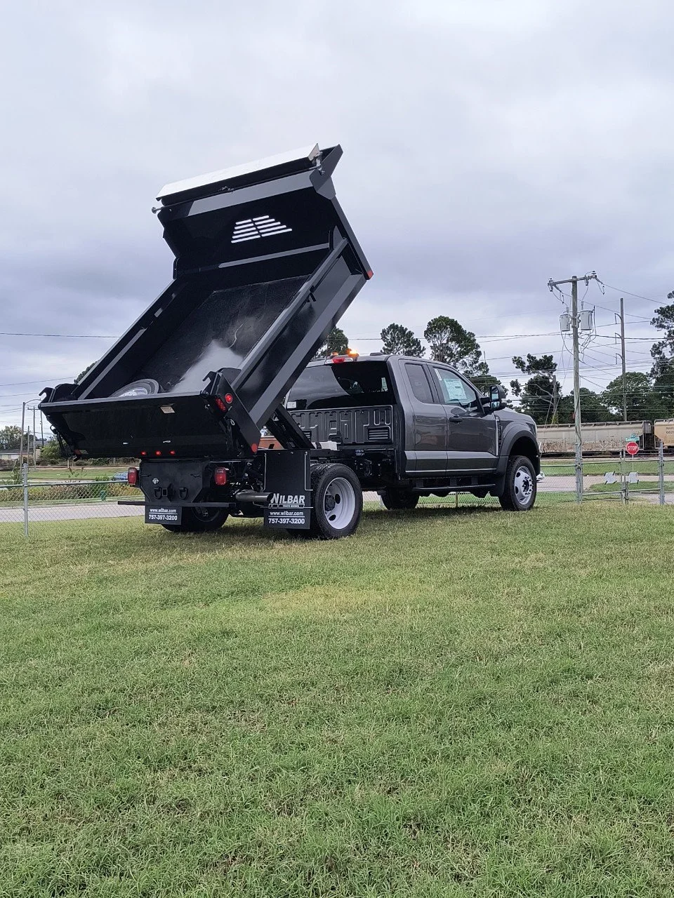 Black utility truck with dump bed raised, parked on grass, overcast sky, power lines and trees in background.