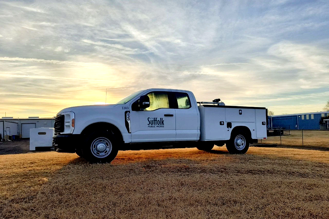 A white Suffolk Public Works utility truck parked on a grassy field at sunset.