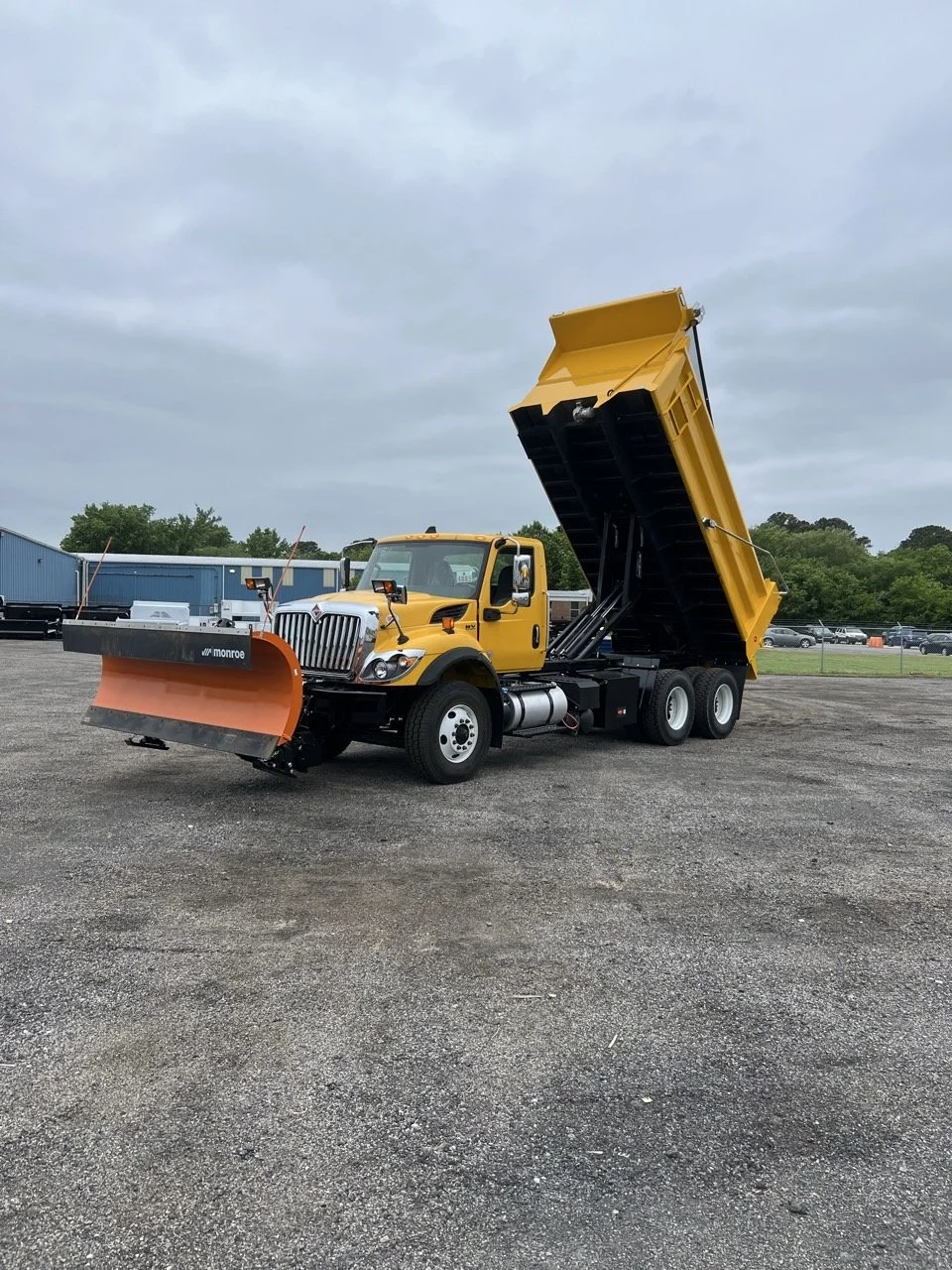 A yellow dump truck with a black and orange snowplow attached to the front and the dump bed raised, parked on a gravel lot under cloudy skies.