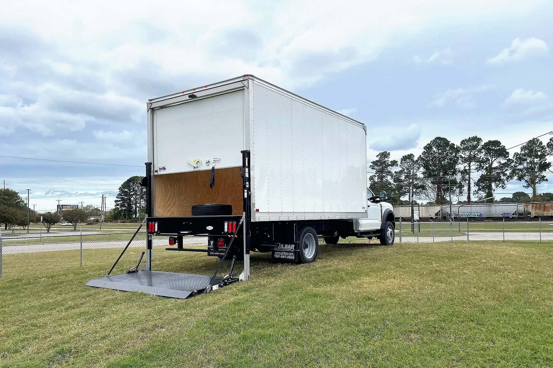 A white box truck with a partially open rear door parked on a grassy field. The truck has a ramp extended onto the ground. In the background, there are trees, a chain-link fence, and other trucks.