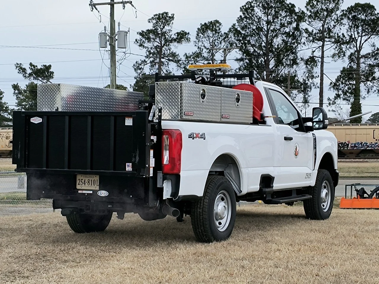 A white utility pickup truck with storage compartments and equipment, parked on a grassy area near train tracks, with trees and power lines in the background.