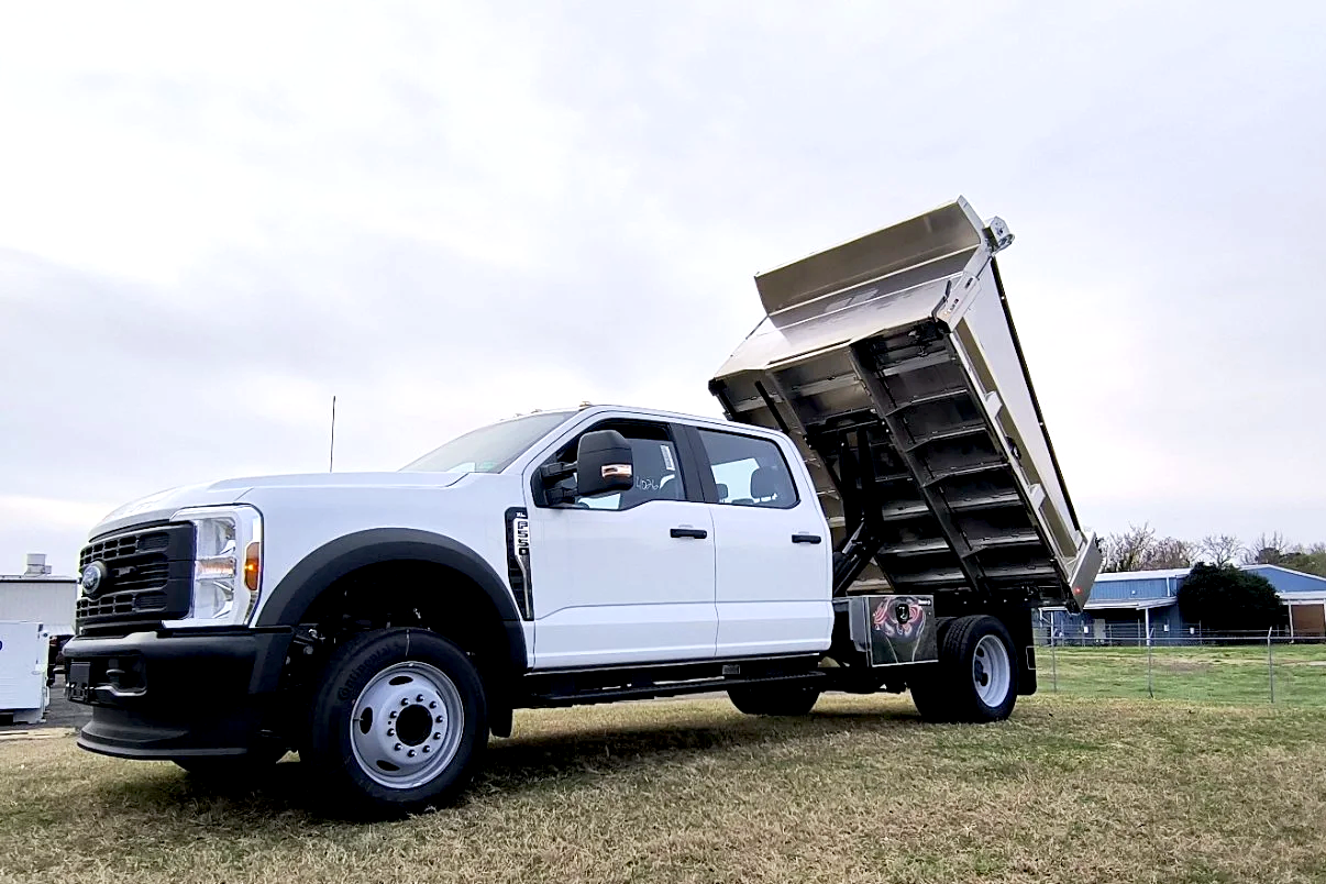 A white dump truck with its bed lifted, parked on grass in a residential or rural area.