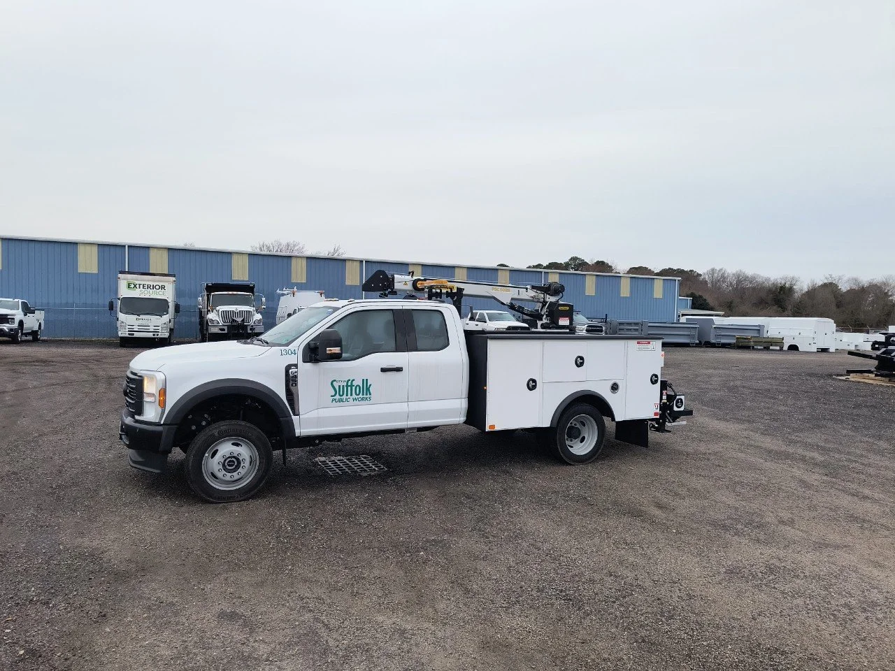 A white Suffolk Public Works utility truck with a crane on the back parked on a gravel lot with other trucks and trailers in the background and a blue industrial building.