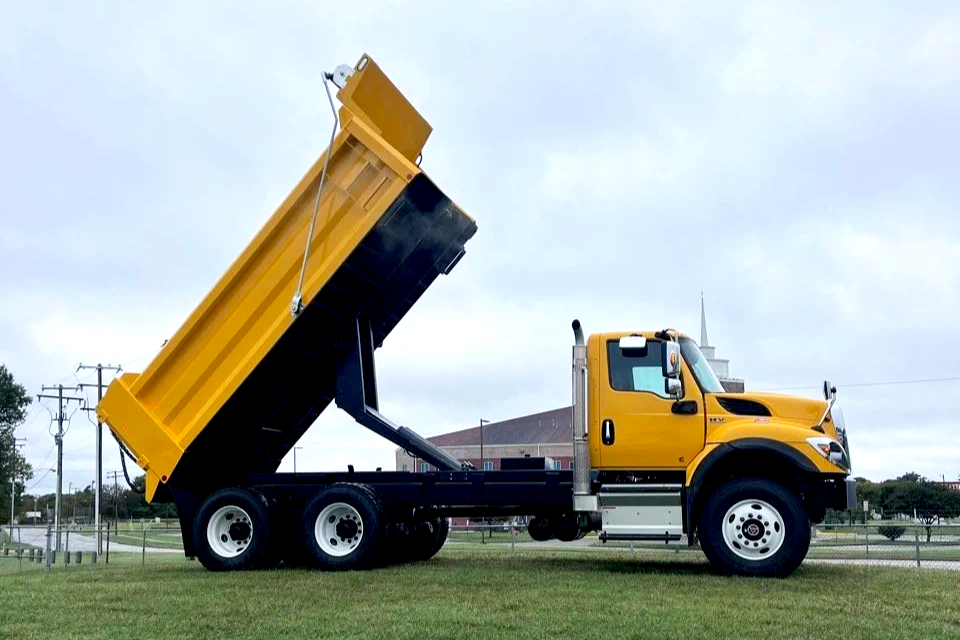 Yellow dump truck with its bed raised, parked on grass with utility poles and buildings in the background.