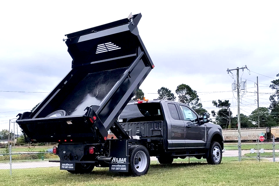 A black dump truck with its bed raised, parked on a grassy area near a fence, electric poles, and a train in the background.