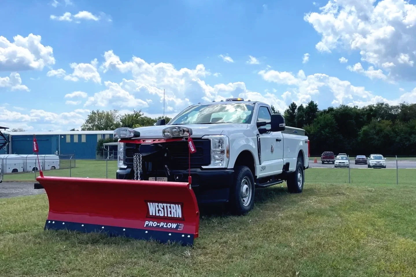 White utility truck fitted with a red Western Pro-Plow 3 snow plow attachment, parked on grassy field with blue sky, clouds, and other vehicles in the background.